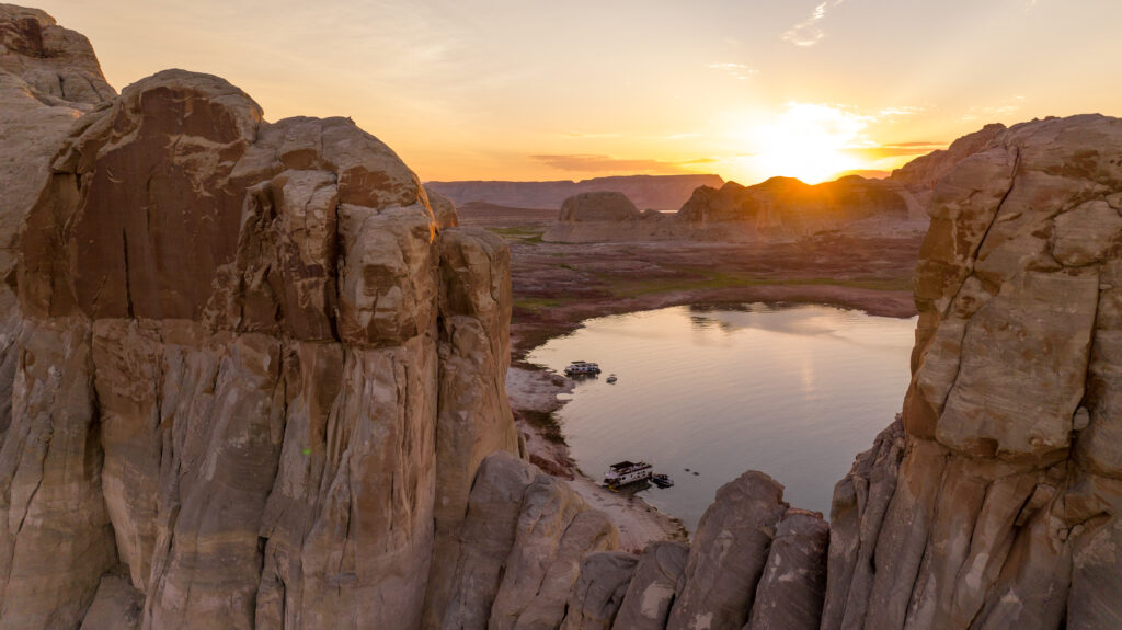 Aerial view of Lake Powell's tall red rock surrounding glassy, reflective water. The sun can be seen setting behind the rocks.
