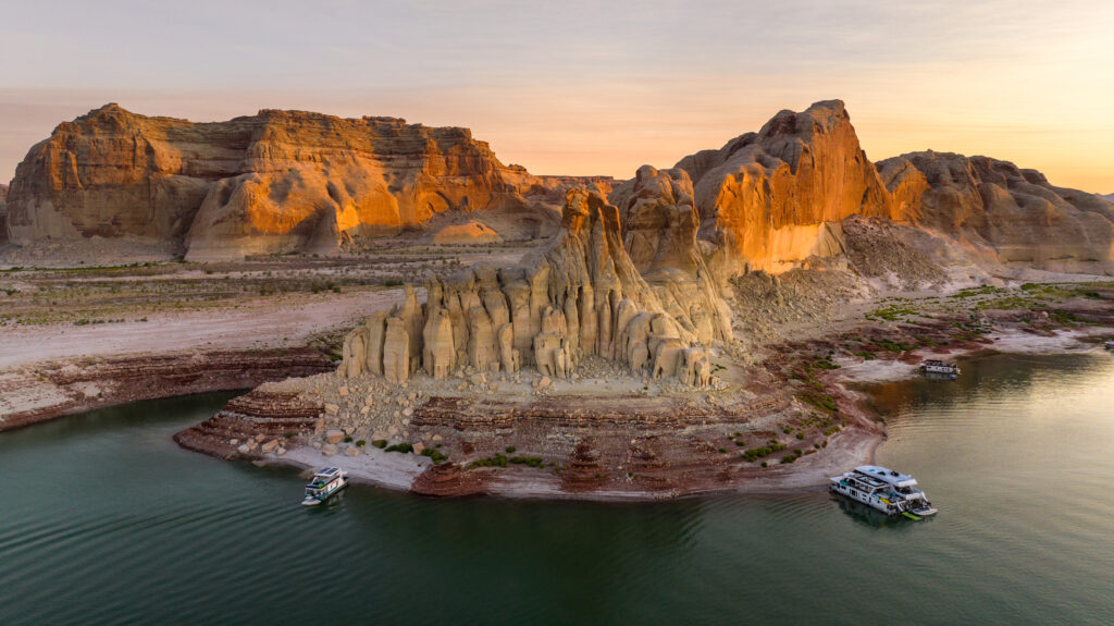Lake Powell aerial view. An expanse of green water and tall red rock in front of a sunset of pastels. A houseboat is parked along the rocky shoreline.