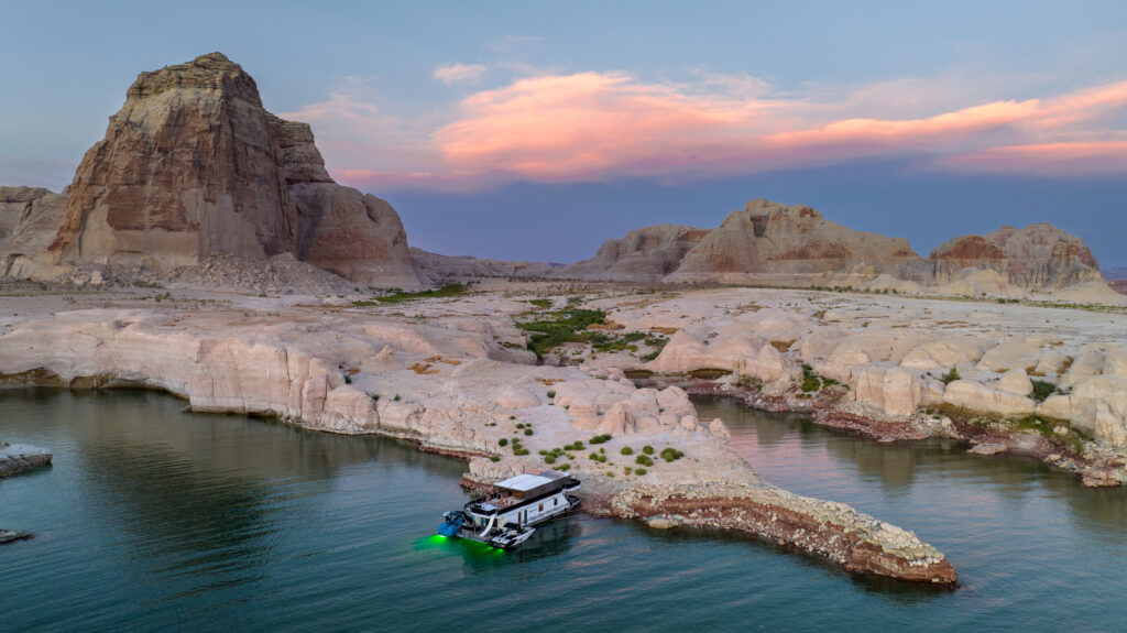 Aerial view of Lake Powell with a cotton candy sunset. Vast, glassy water sits in the foreground and textured red rock stands tall and large in the background. A houseboat equipped with Liquid Lumens underwater boat lights sits on the shoreline.